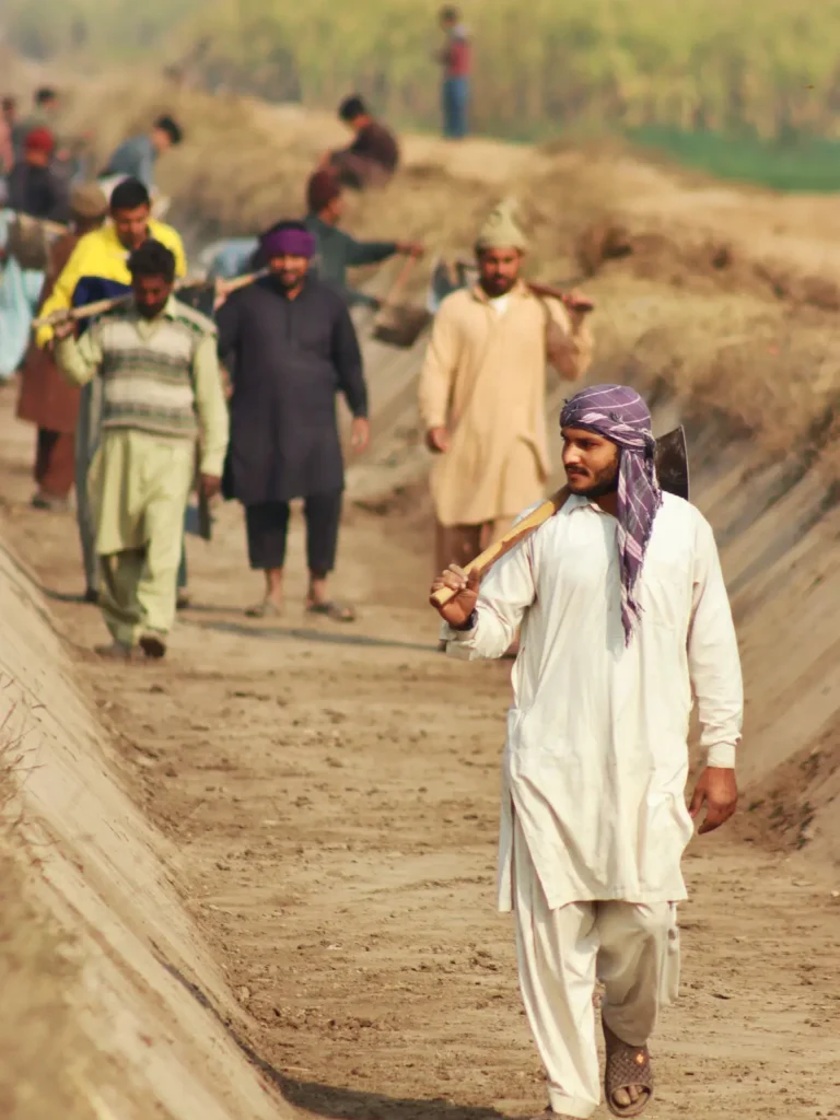 Farmers walk along a dry irrigation channel with tools over their shoulders.