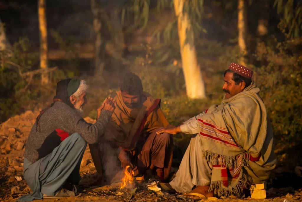 Three farmers sit and talk around a small fire at dusk.