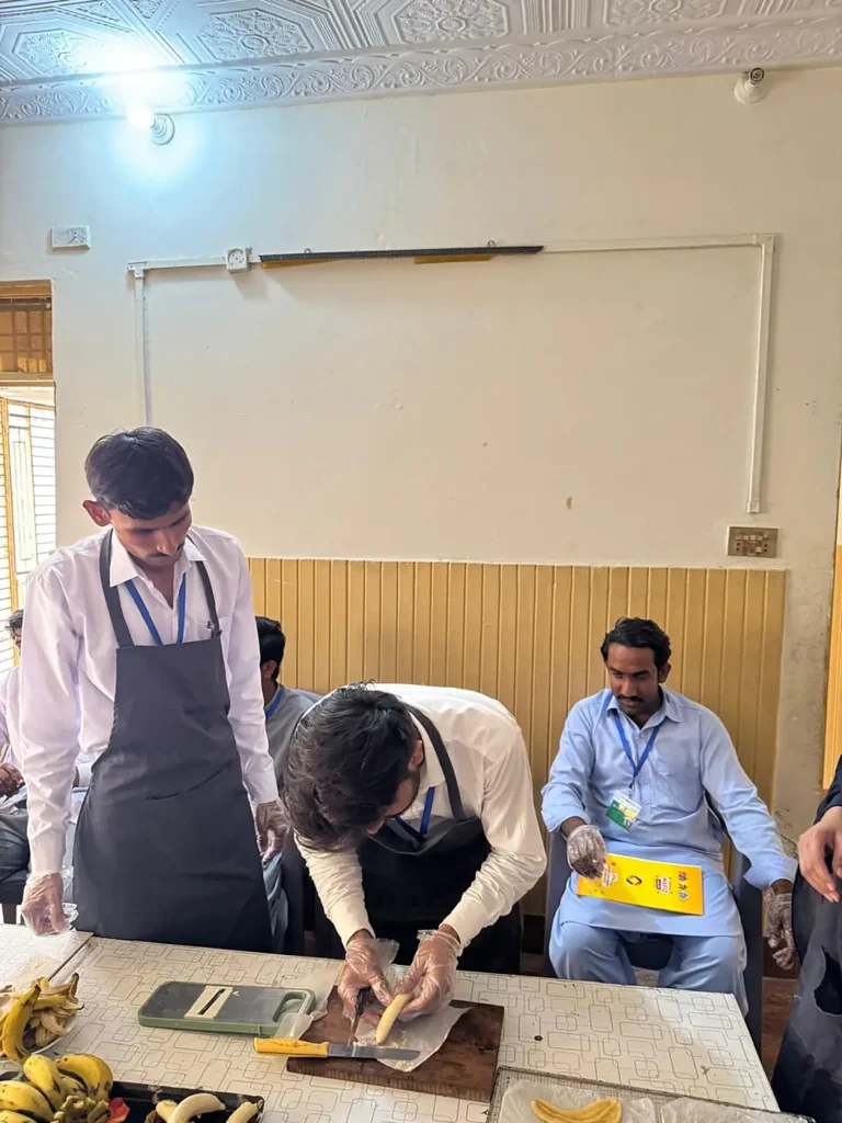Men in aprons and gloves slice bananas at a work table while another man looks on.
