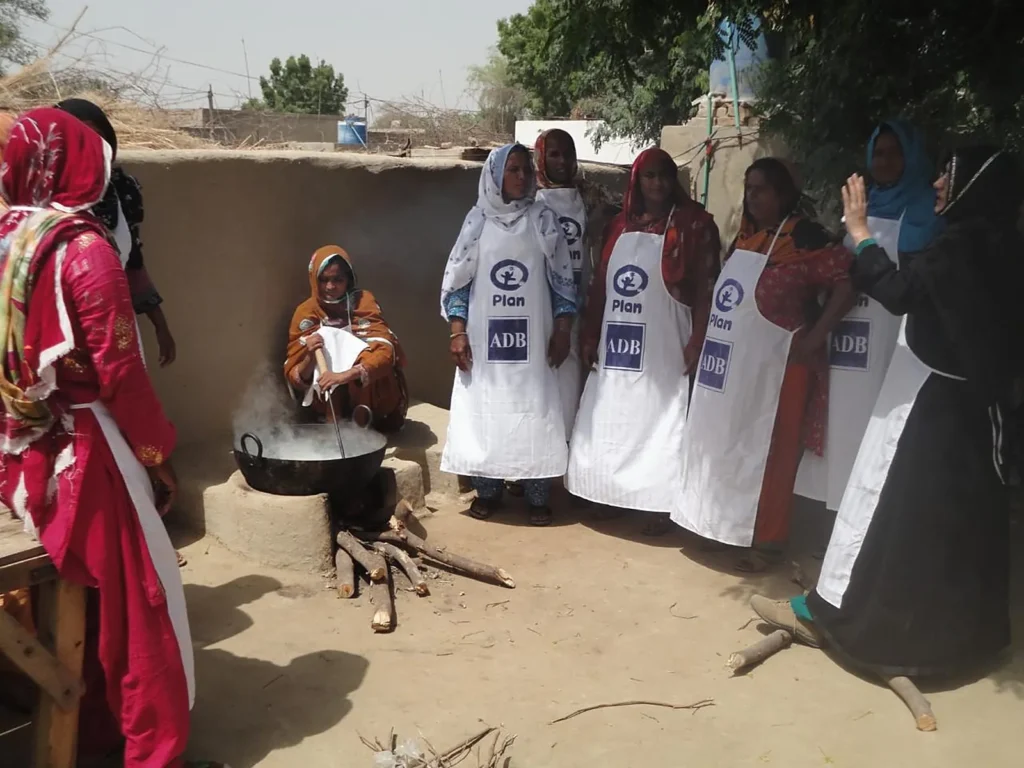 Women in aprons gather in a village courtyard around a large pot for a dairy processing demonstration.