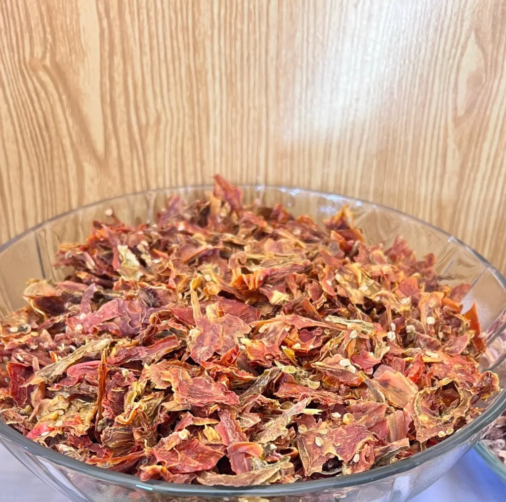 A glass bowl filled with dried red vegetable pieces in front of a wooden panel.