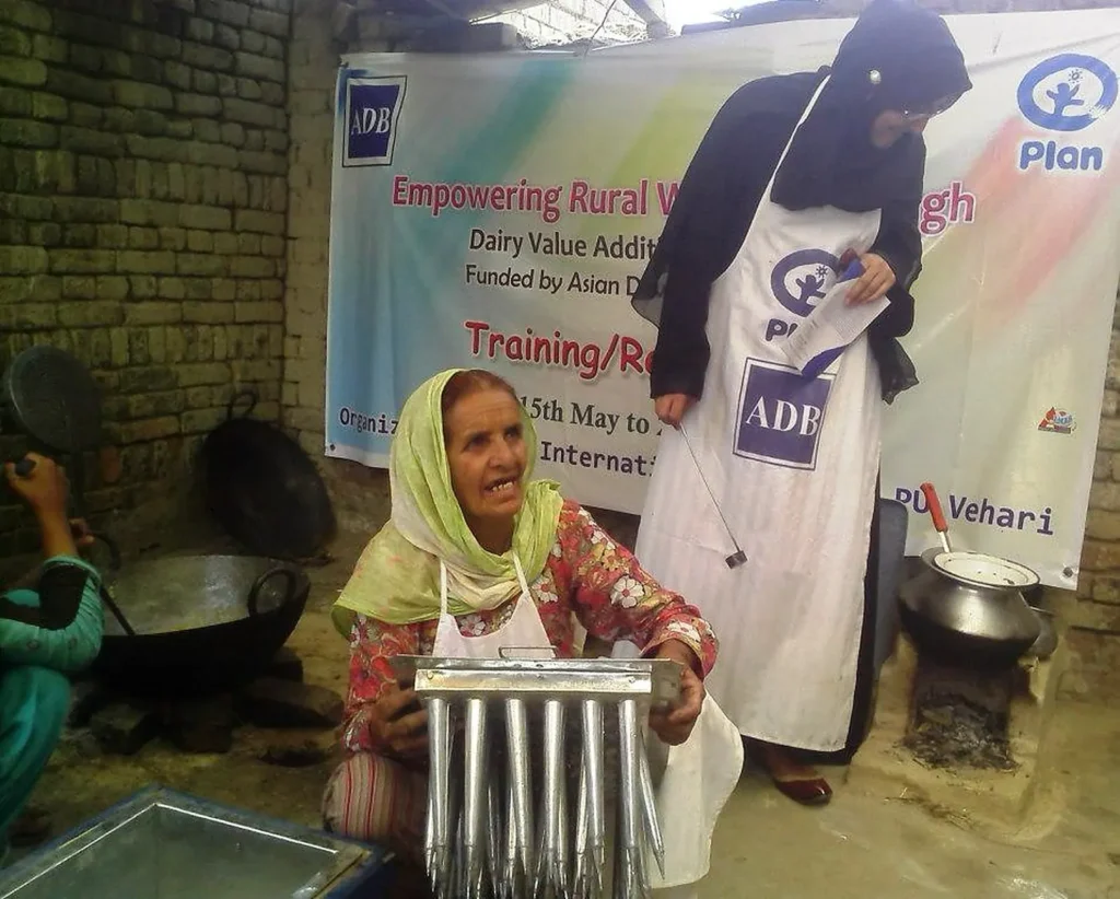 A woman holds a steel mould with rows of tubes while a trainer in an apron looks on at a dairy value-addition session.