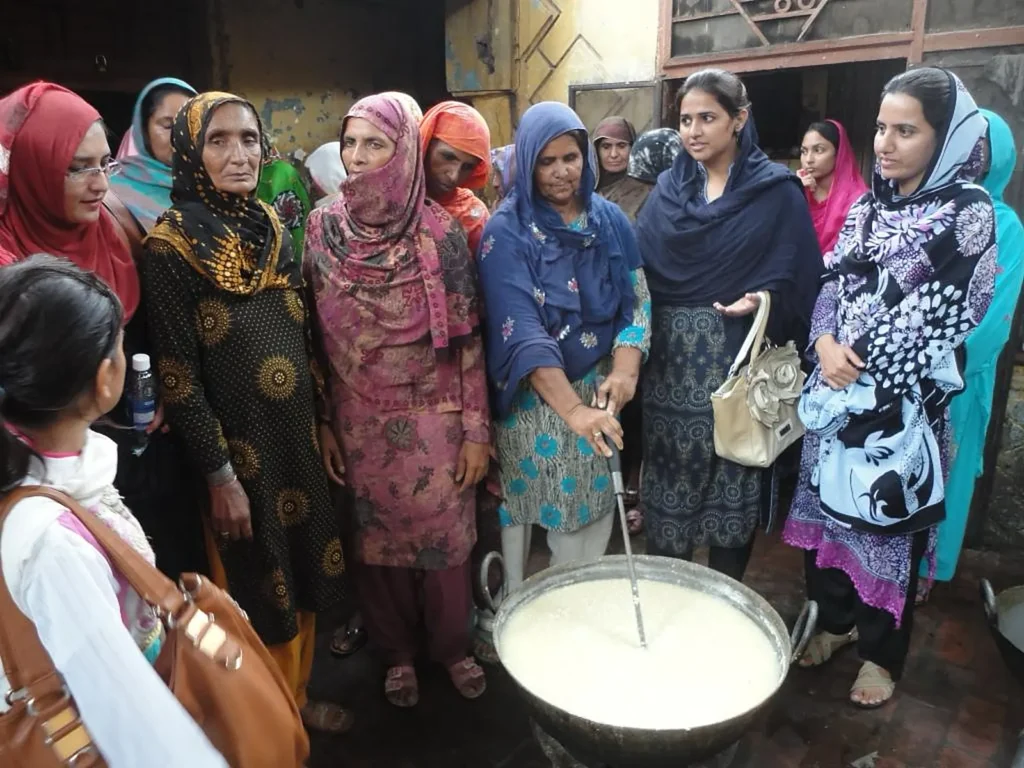 Women gather around a large steel pot of fresh milk during a hands-on dairy processing session.