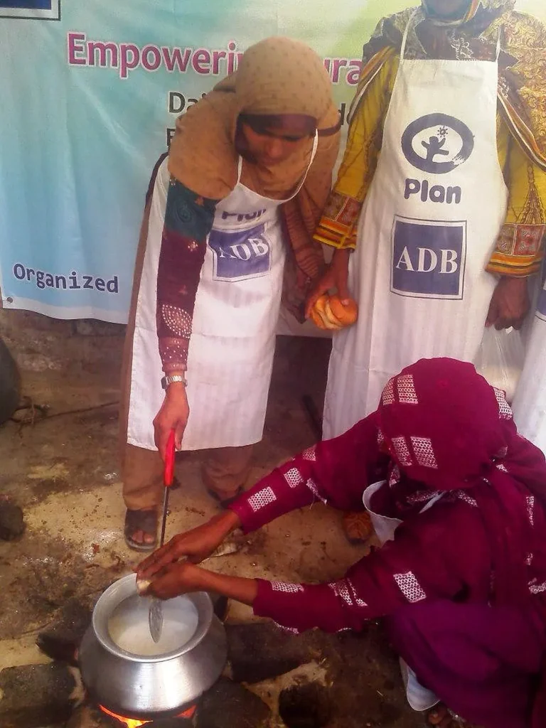Women in aprons stir milk in a steel pot over a fire during a hands-on dairy processing session.