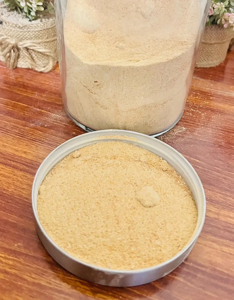 A tall glass jar and a round tin filled with fine light-brown powder on a wooden table.