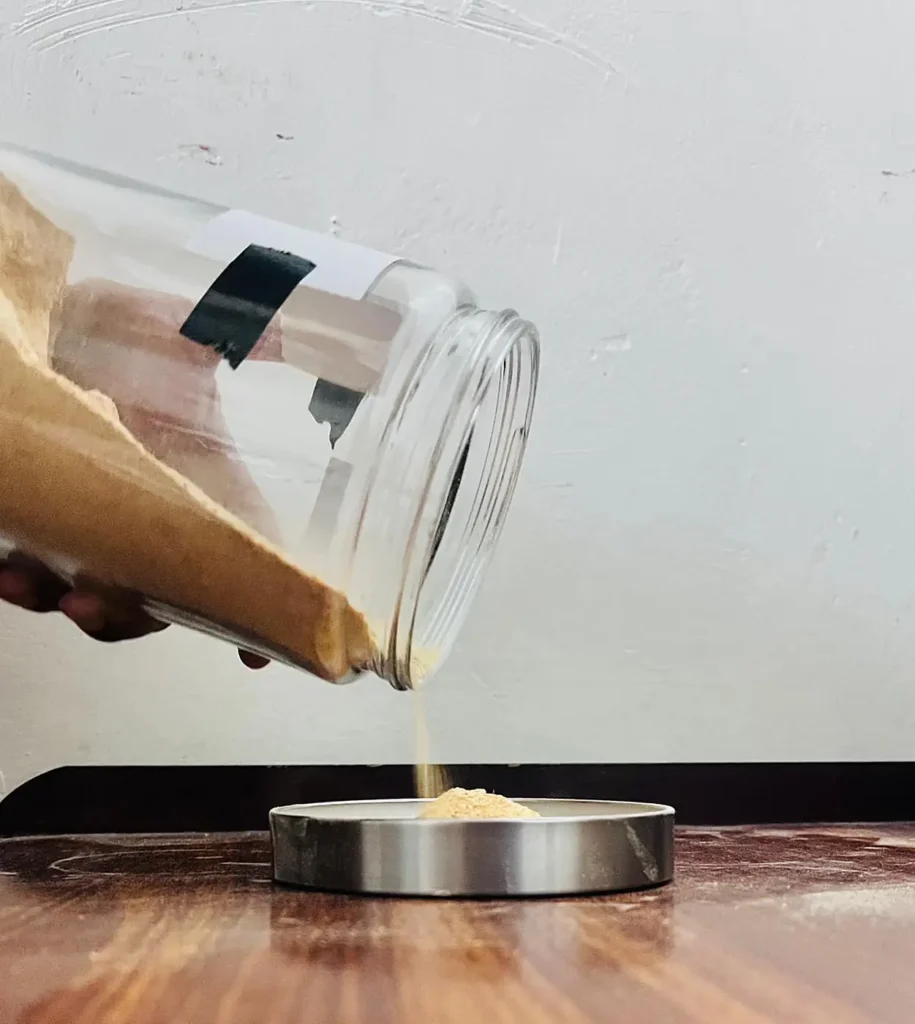 Fine powder pours from a tipped glass jar into a steel tin.