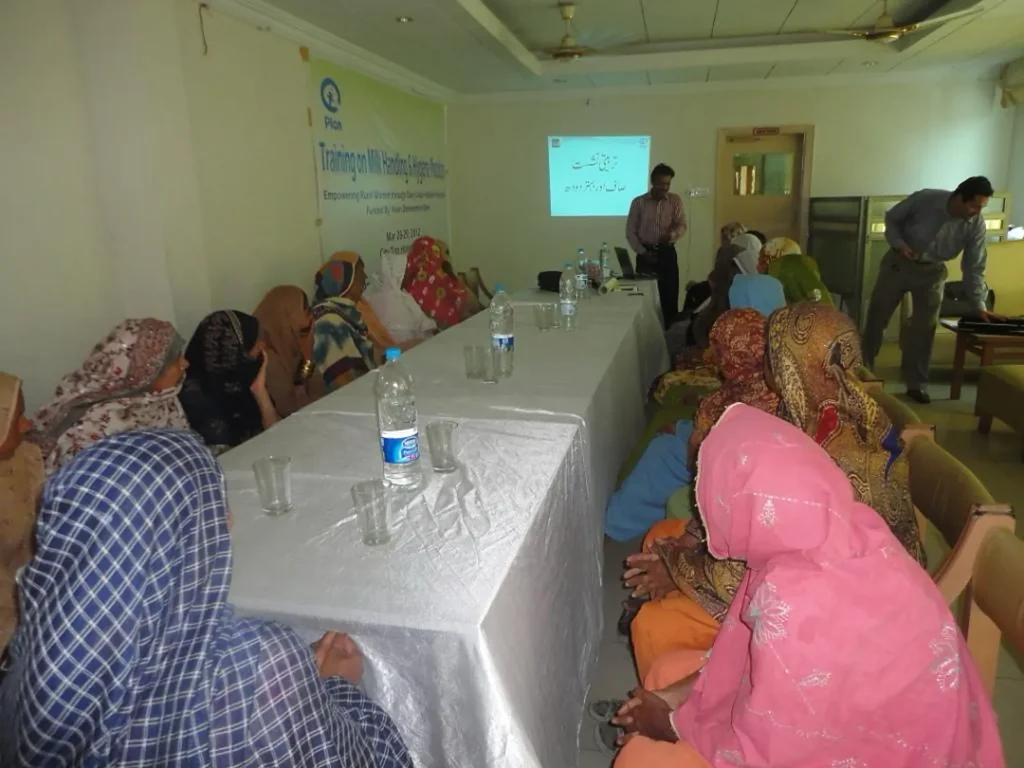 Women trainees sit along a table in a training hall while a trainer presents at the front.