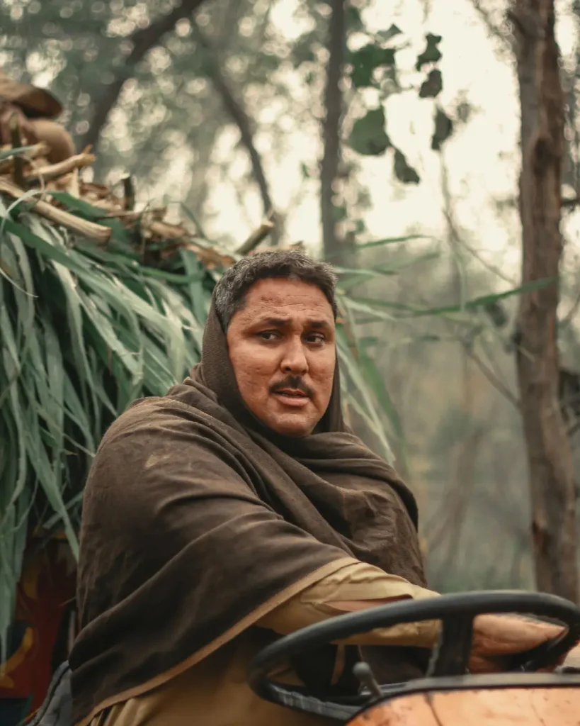 A farmer at the wheel of a tractor beside a heap of green fodder.