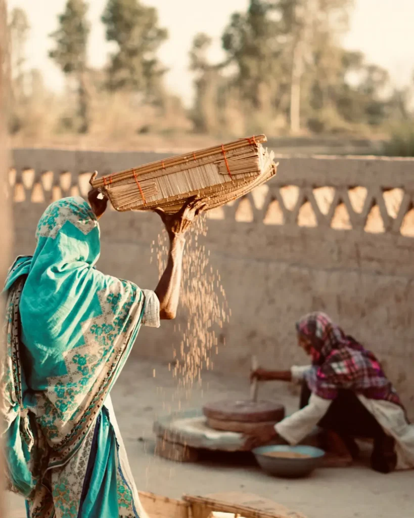 A woman in a turquoise shawl winnows grain with a woven tray beside a patterned mud wall.